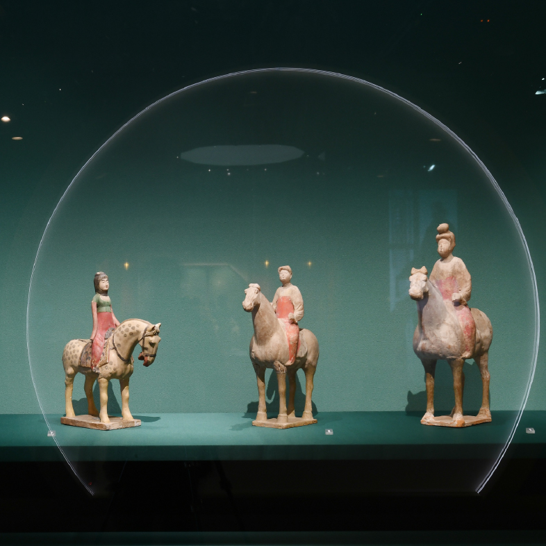 Four ancient Chinese ceramic figurines of riders on horseback, displayed in a row inside a museum exhibit.